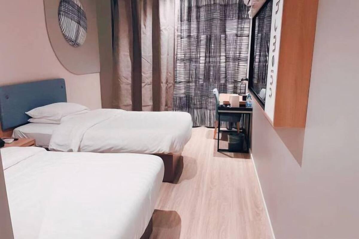 A bedroom with two beds, a bedside table, a small desk with a chair, and a window with curtains.