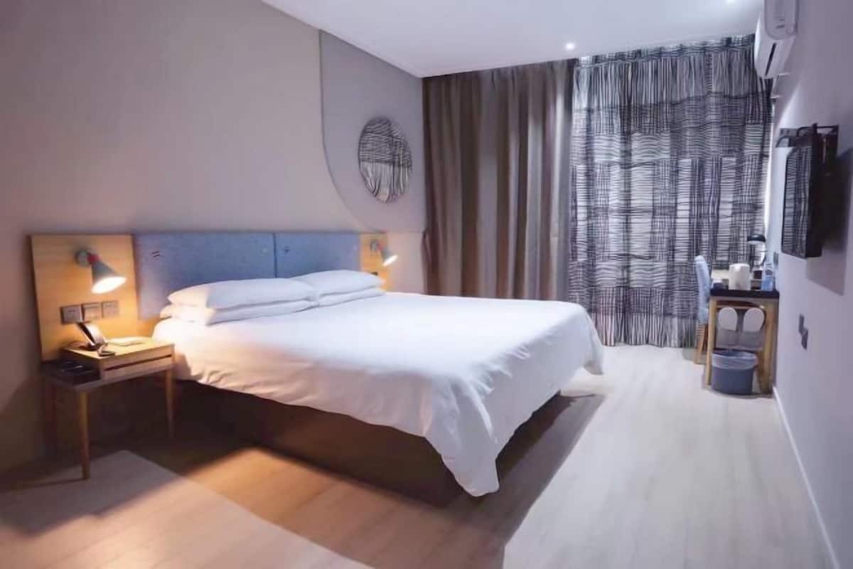 A neatly made bed in a hotel room with a headboard, bedside lamps, a wall-mounted TV, and a curtain.