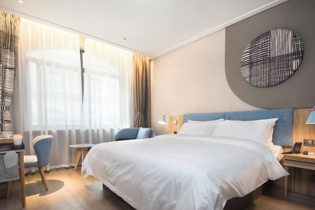 A hotel room with a large bed, a desk with a chair, a round wall decoration, and a view of the outdoors through the window.