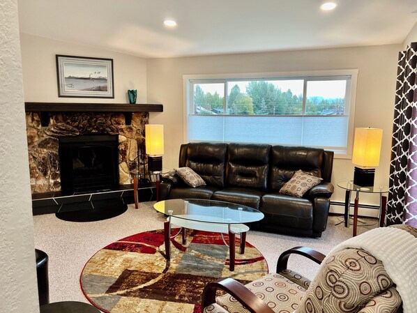 A living room with a fireplace, a leather sofa, a glass coffee table, and a view of the outdoors. - NEW LISTING! Adventure and Unwind:View of Mountains, Near AirP, Lake w 2 Kayaks (Anchorage)