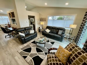 A modern living room with two black leather sofas, a glass coffee table, and a patterned rug. There is a dining area with a table and chairs, and a kitchen area with a microwave and stove. - NEW LISTING! Adventure and Unwind:View of Mountains, Near AirP, Lake w 2 Kayaks (Anchorage)