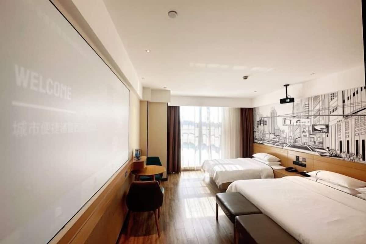 Habitación de hotel con dos camas, un escritorio, una silla, una mesita y una pantalla de proyección que muestra 'WELCOME'.