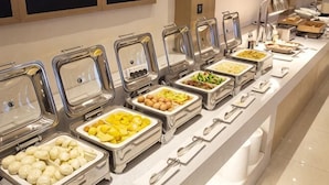 Una barra de buffet con distintos alimentos en recipientes metálicos, un mostrador blanco y una lámpara fijada a la pared.