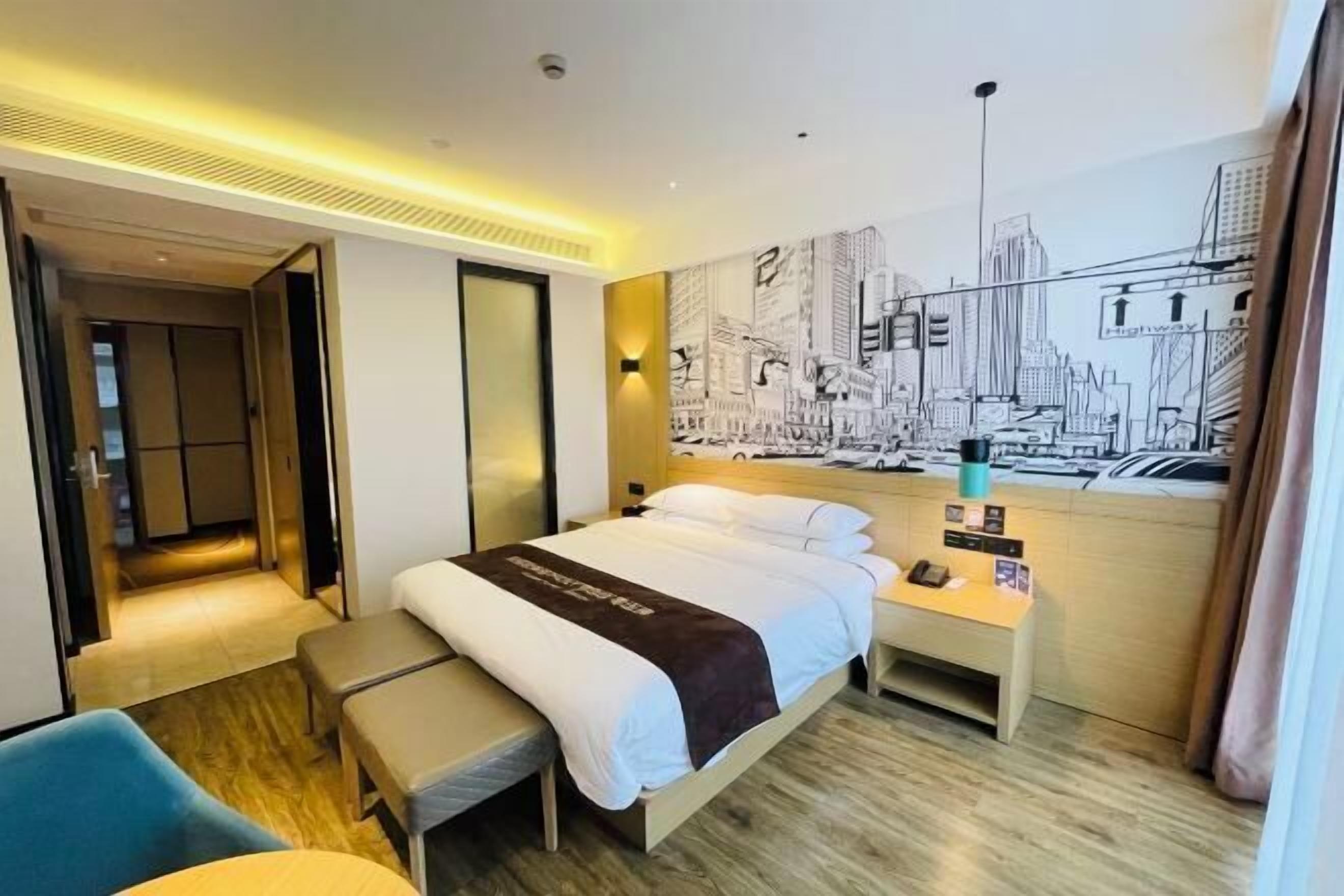 A hotel room with a bed, bedside table, bench, and a cityscape mural on the wall.