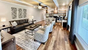 A modern living room with a sofa, coffee table, and armchairs, a kitchen with wooden cabinets and appliances, and a large window with blinds. - Mountainside Luxury Unit 3—LOCATION! LOCATION! (Mena)