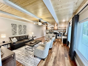 A modern living room with a sofa, coffee table, and armchairs, a kitchen with wooden cabinets and appliances, and a large window with blinds. - Mountainside Luxury Unit 3—LOCATION! LOCATION! (Mena)