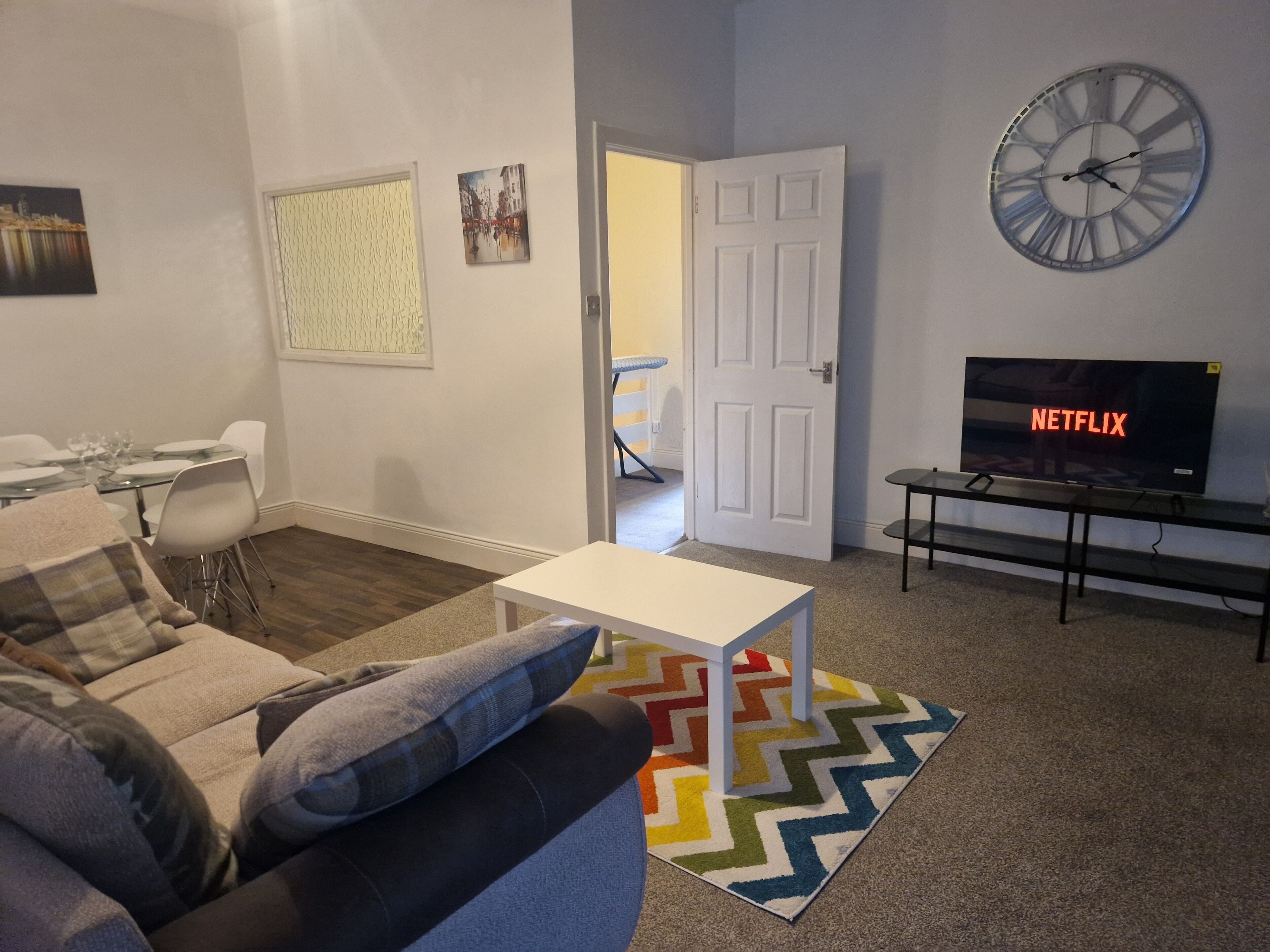 A living room with a sofa, a small table, a TV displaying 'NETFLIX', a clock, and a colorful rug.