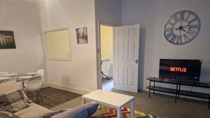 En stue med en sofa, et lille bord, et fjernsyn der viser 'NETFLIX', et ur og en farverig måtte.