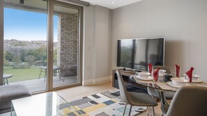 En moderne stue med et stort fjernsyn, et spisebord dækket til fire personer, og en skydedør, der fører ud til en balkon med udsigt over byen.