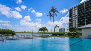 Uma piscina com água azul cristalina, rodeada por palmeiras e um edifício moderno com fachada de vidro.