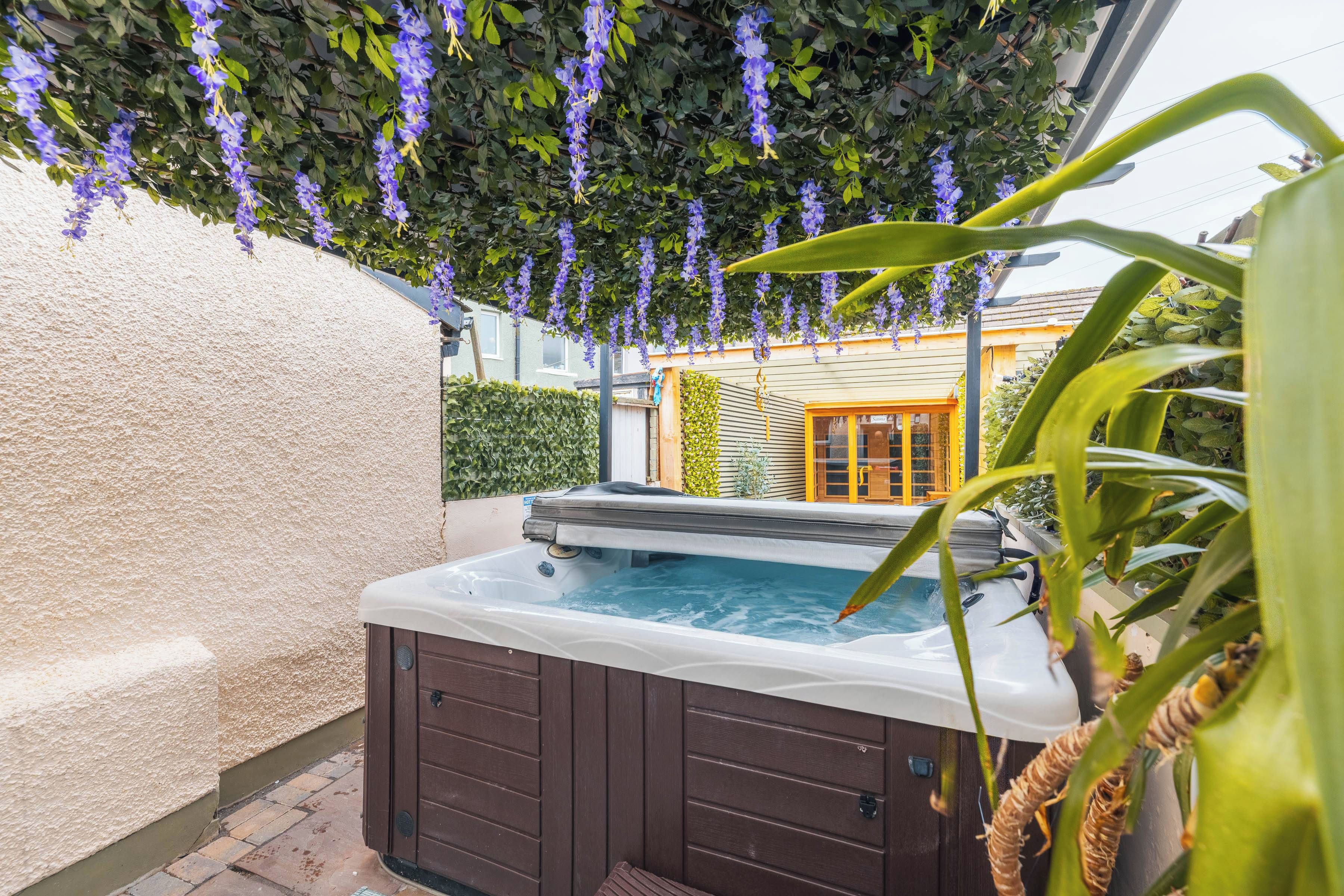 A hot tub with a wooden exterior, surrounded by a wall and a garden with hanging purple flowers.