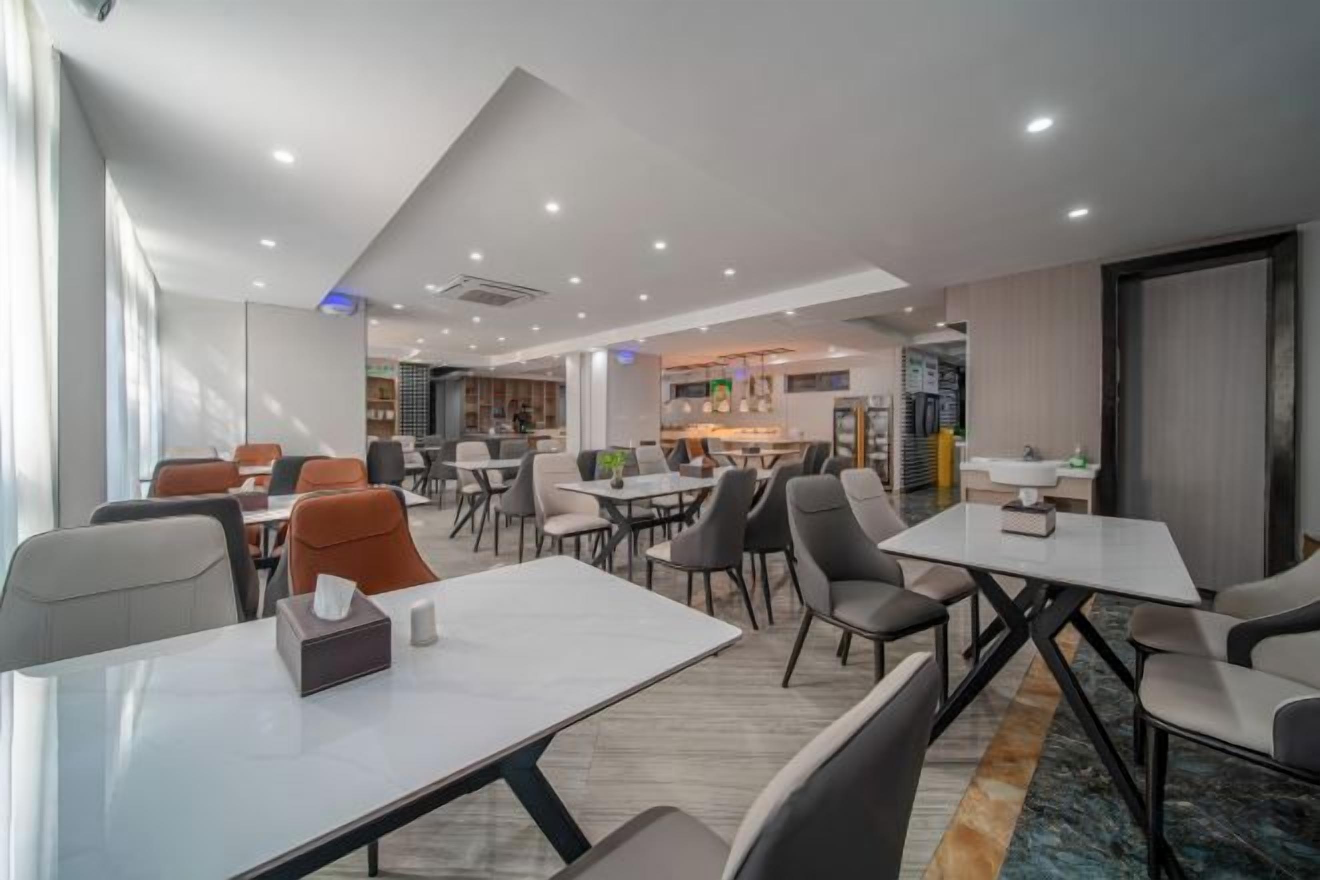 A modern hotel dining area with multiple tables and chairs, a kitchen in the background, and a large window providing natural light.