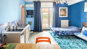 Ein gemütlicher Raum mit Küchenzeile, Essbereich, einem Bett mit blau gemustertem Überwurf und einem großen Fenster mit Vorhängen.