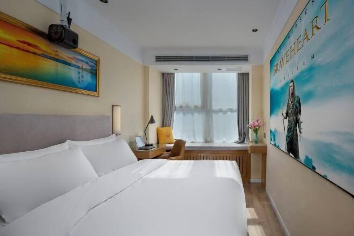 Una habitación de hotel con una cama grande, un escritorio con lámpara, una silla, una ventana con cortinas y una pared con un cartel de película.