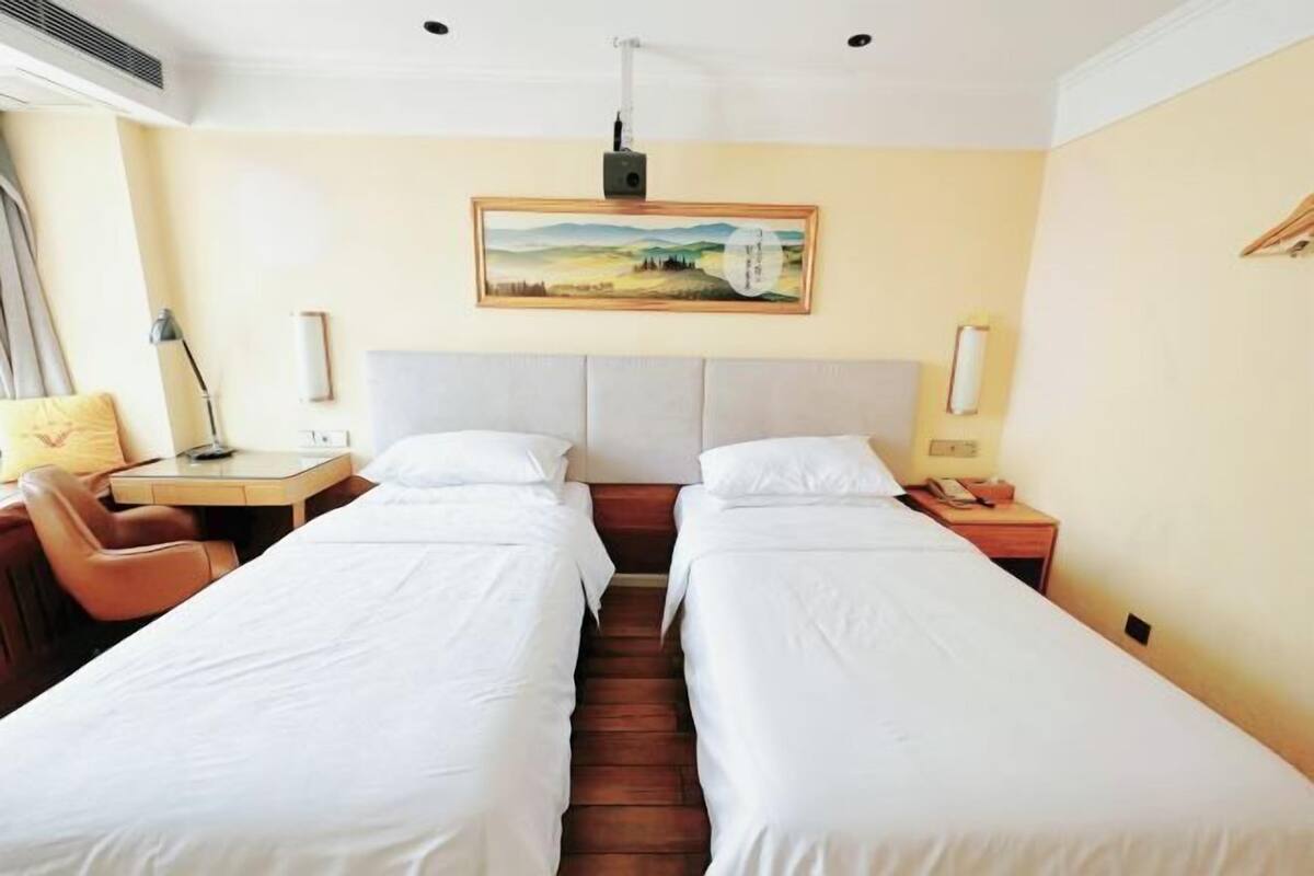 Habitación de hotel con dos camas individuales, un escritorio, una silla, una mesita de noche, una lámpara y un cuadro en la pared.