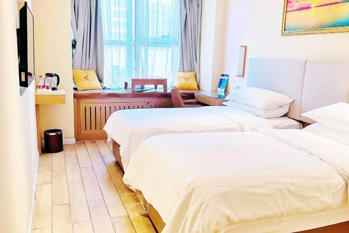 Una habitación de hotel con dos camas, suelo de madera, ventana con cortinas, una mesita con un ordenador portátil y un cuadro en la pared.