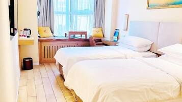 A hotel room with two beds, a wooden floor, a window with curtains, a small table with a laptop, and a painting on the wall.
