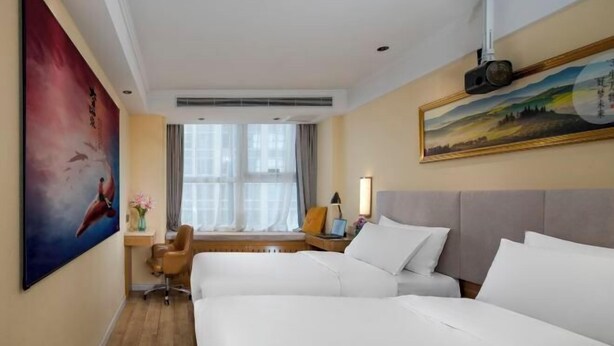 Habitación de hotel con dos camas, un escritorio, una silla, una ventana con cortinas y obras de arte en las paredes.