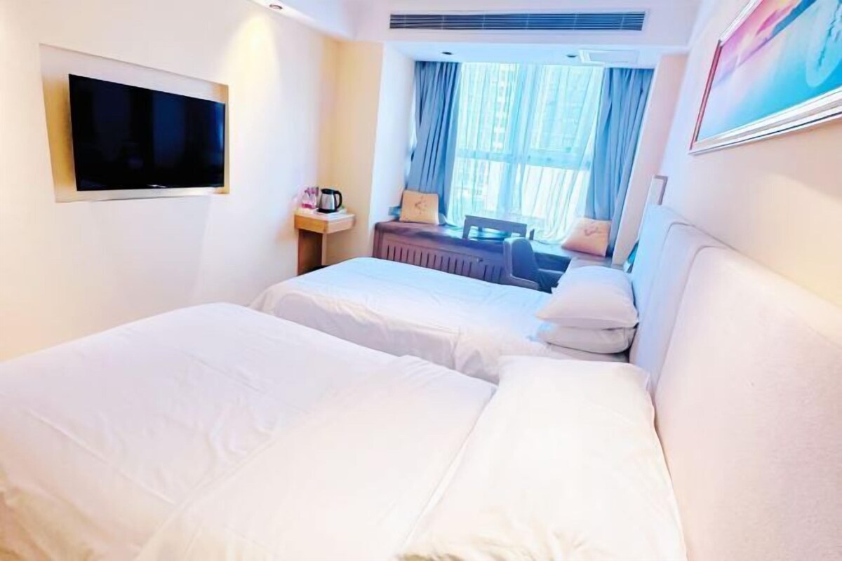 A hotel room with two beds, a flat-screen TV mounted on the wall, a small table with a lamp, and a window with curtains.
