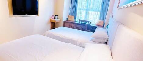 A hotel room with two beds, a flat-screen TV mounted on the wall, a small table with a lamp, and a window with curtains.