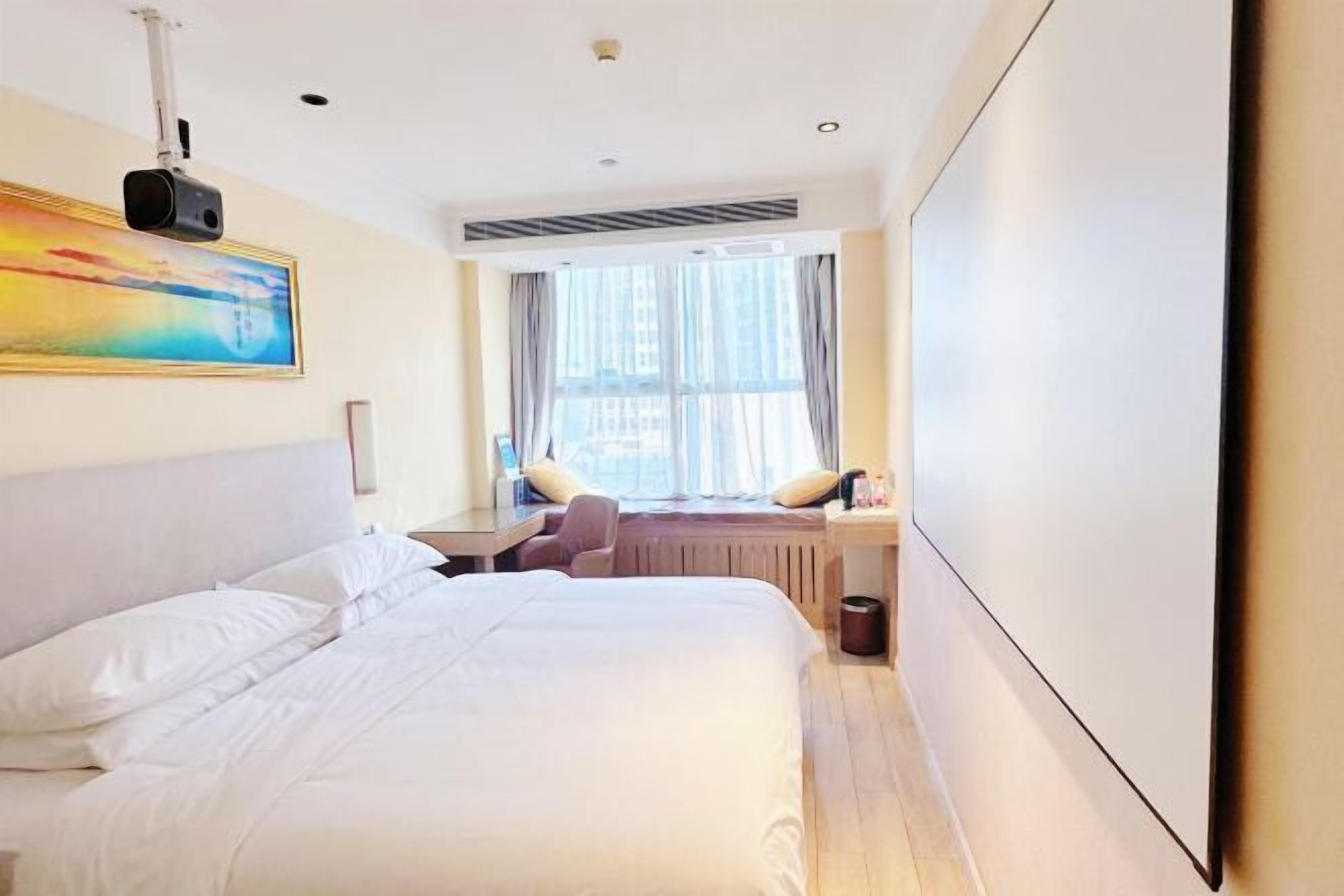 A hotel room with a bed, a desk, a chair, a window with curtains, and a painting on the wall.