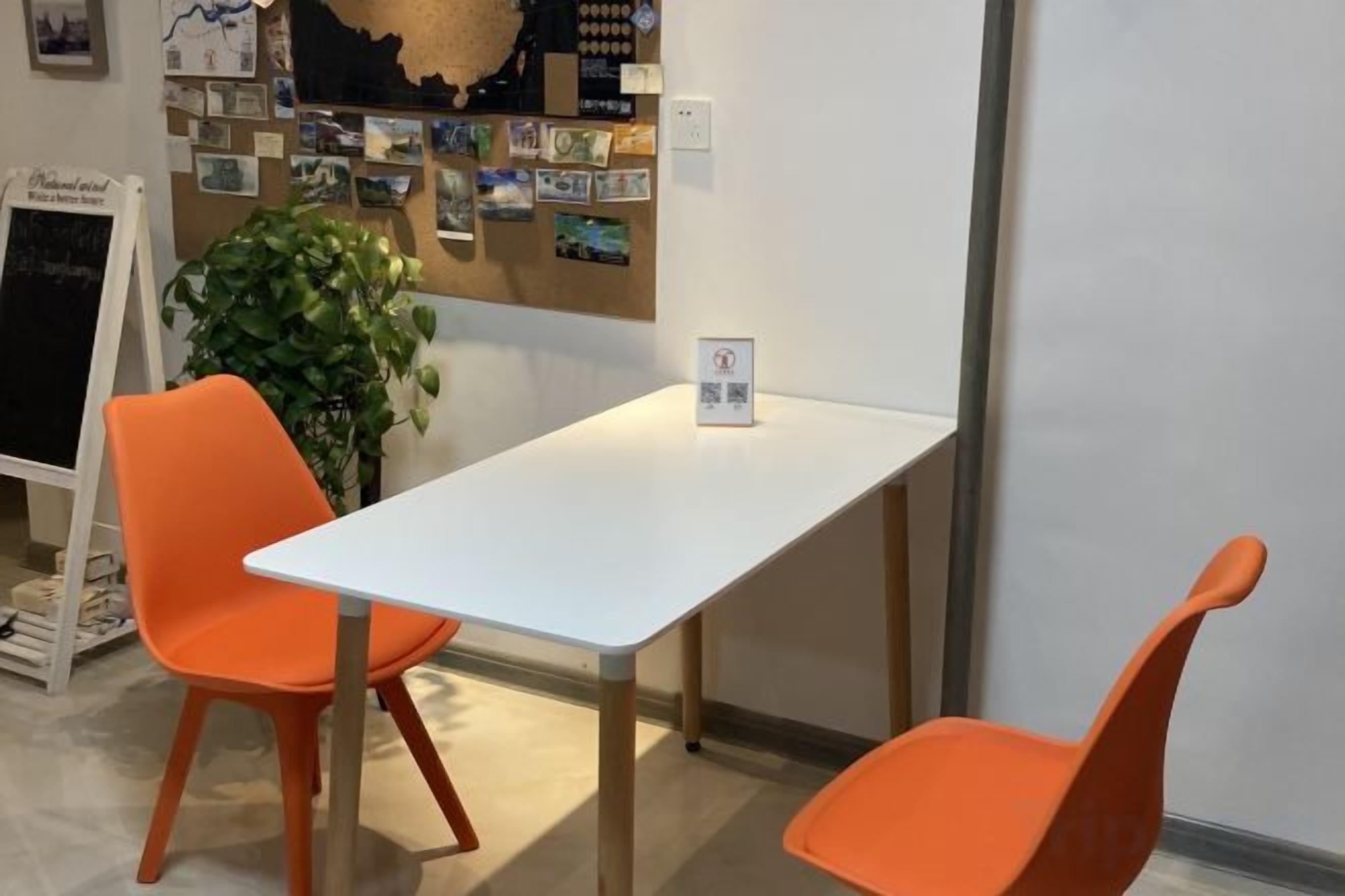 A room with a white table and two orange chairs, a bulletin board with photos, and a plant.