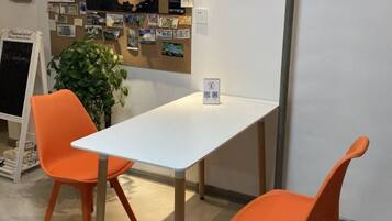 A room with a white table and two orange chairs, a bulletin board with photos, and a plant.