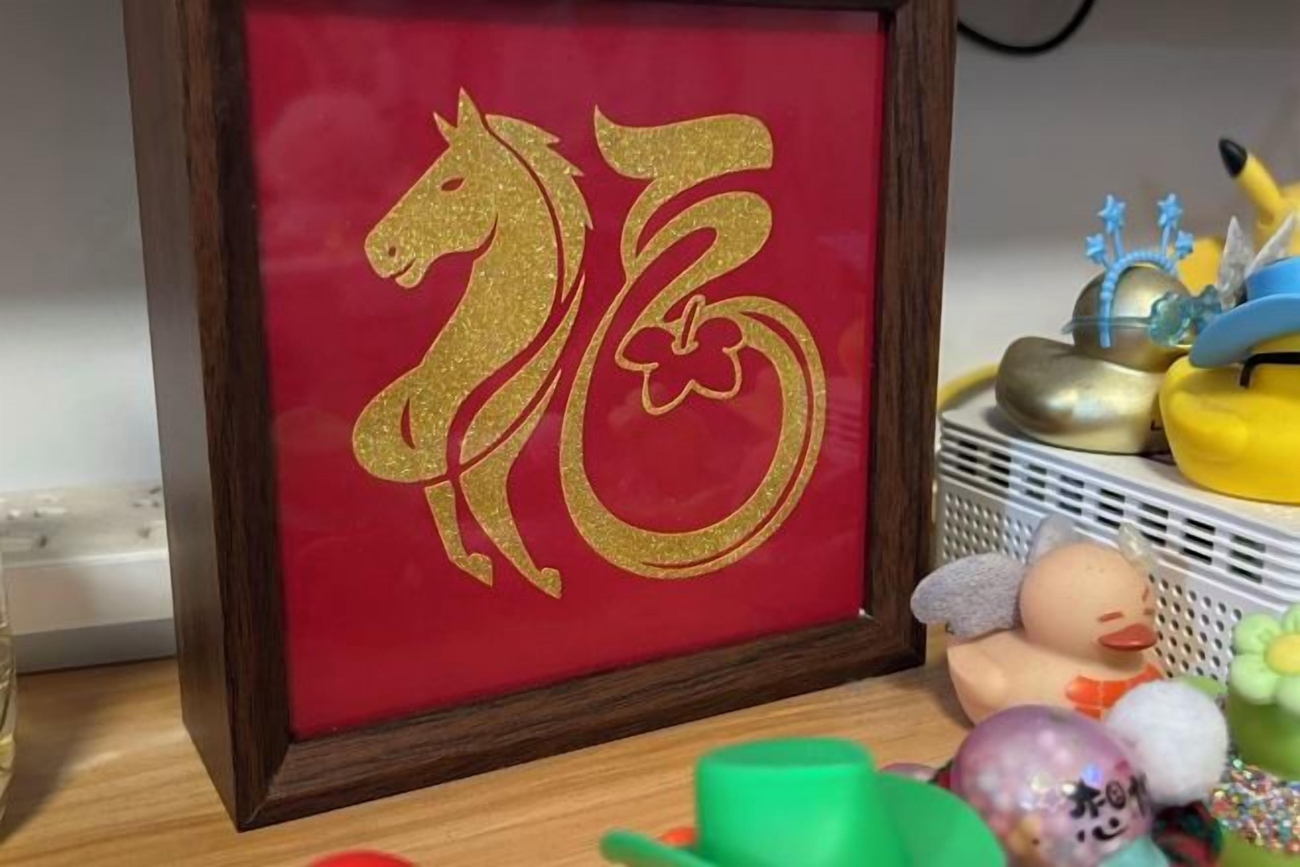 Un'opera d'arte incorniciata raffigurante un cavallo dorato su sfondo rosso, un cesto bianco con diversi oggetti e un oggetto verde su un tavolo.