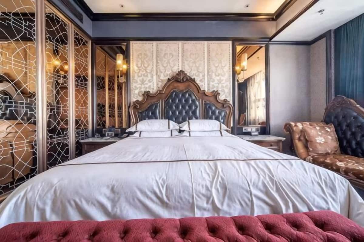 A large bed with a wooden headboard, a nightstand, and a chair in a room with patterned wallpaper and a decorative wall.