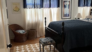 Un dormitorio con cama, una silla, un taburete pequeño, un ventilador de techo y una alfombra estampada.