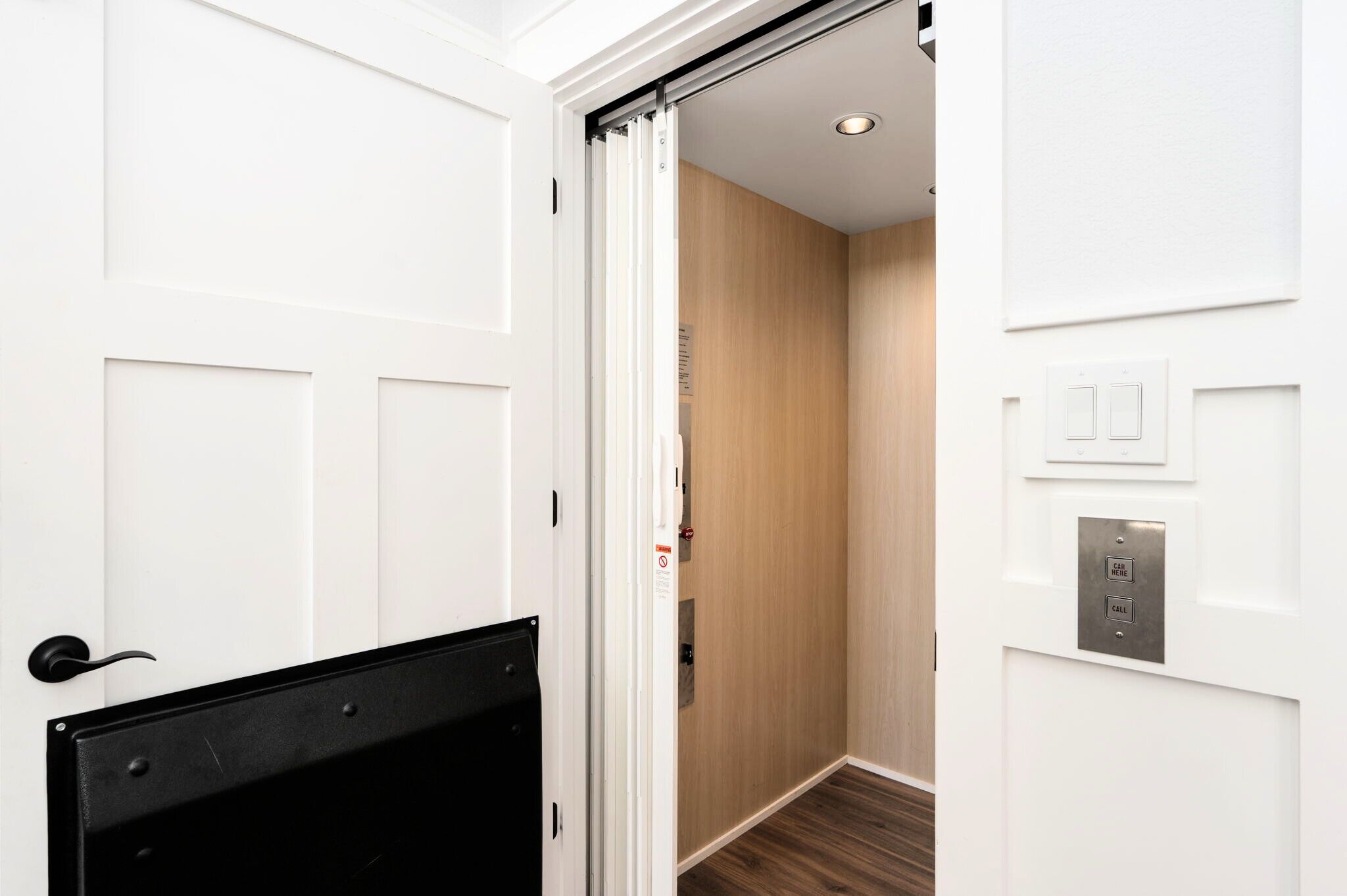 Habitación de hotel con puerta corrediza que da a un pasillo con piso de madera y un botón de llamada fijado a la pared.