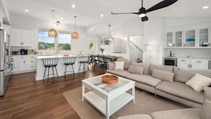 A modern kitchen with white cabinets, a central island with bar stools, a ceiling fan, and a dining area with a staircase. - Last-Minute! Soundfront, Pool, Hot Tub, Elevator, Renowned Currituck Club Access (Corolla)