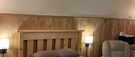 A wooden-paneled bedroom with a bed, bedside lamps, a small table, and a chair.