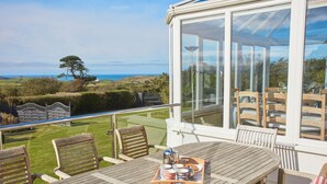 Une terrasse en bois avec une table et des chaises, un espace clos par des baies vitrées offrant une vue sur la campagne, et un plateau avec divers objets posés sur la table.