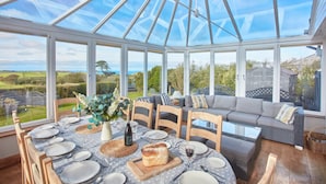 Une véranda avec une table à manger dressée pour six personnes, entourée de chaises en bois, d’un canapé et d’une table basse en verre.
