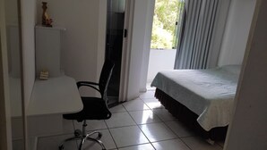 A bedroom with a bed, a desk with a chair, a small table with a decorative item, and a sliding glass door leading to a balcony. - Itapema - Ap. Inteiro em Itapema Rua 129 B (Centro Itapema)