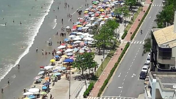 A beach with umbrellas and people, a road with cars, and buildings in the background. - Itapema - Ap. Inteiro em Itapema Rua 129 B (Centro Itapema)