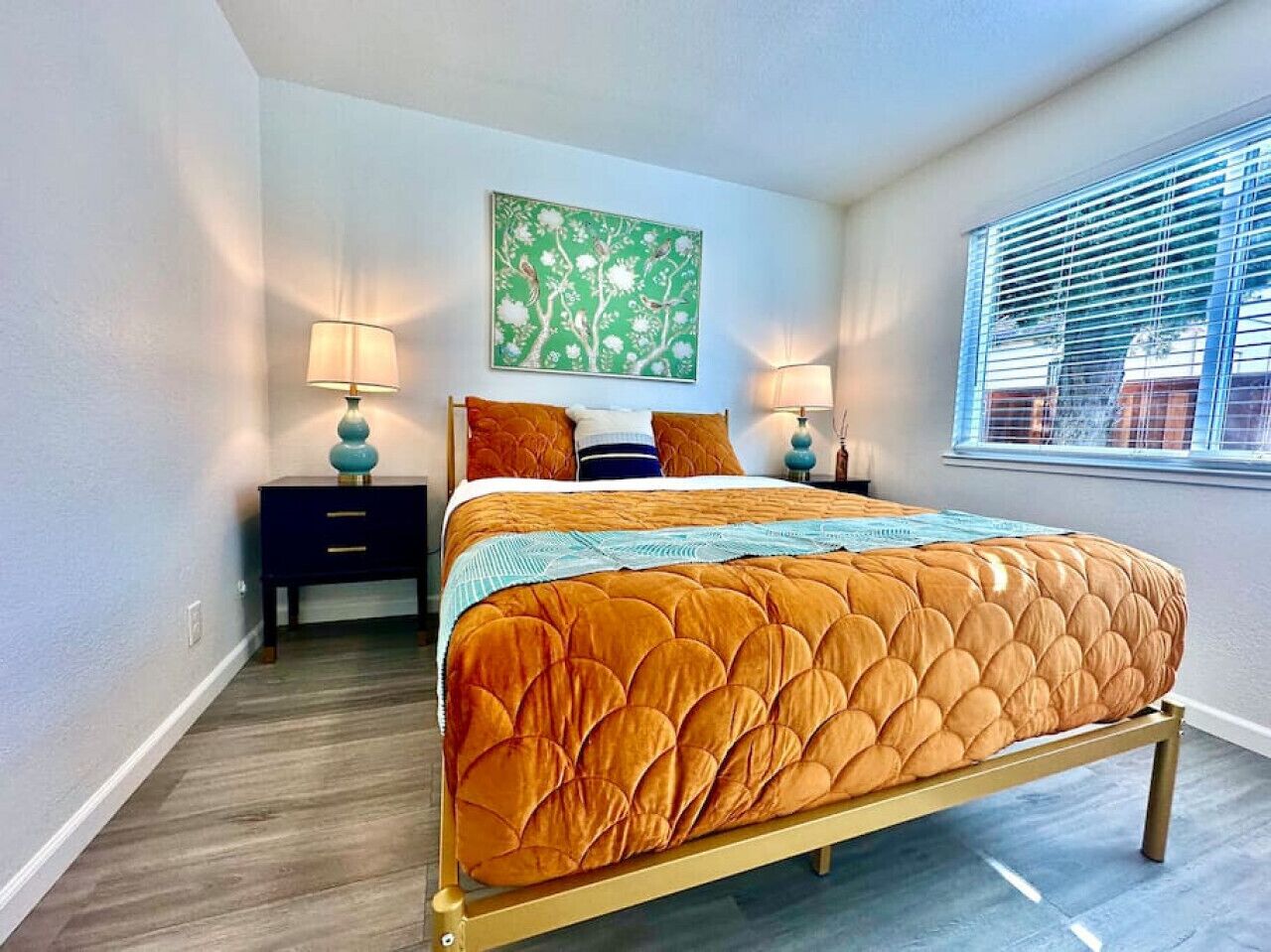 Un dormitorio con cama, dos mesitas de noche, una mesita auxiliar, una ventana con persianas y un cuadro abstracto verde en la pared.