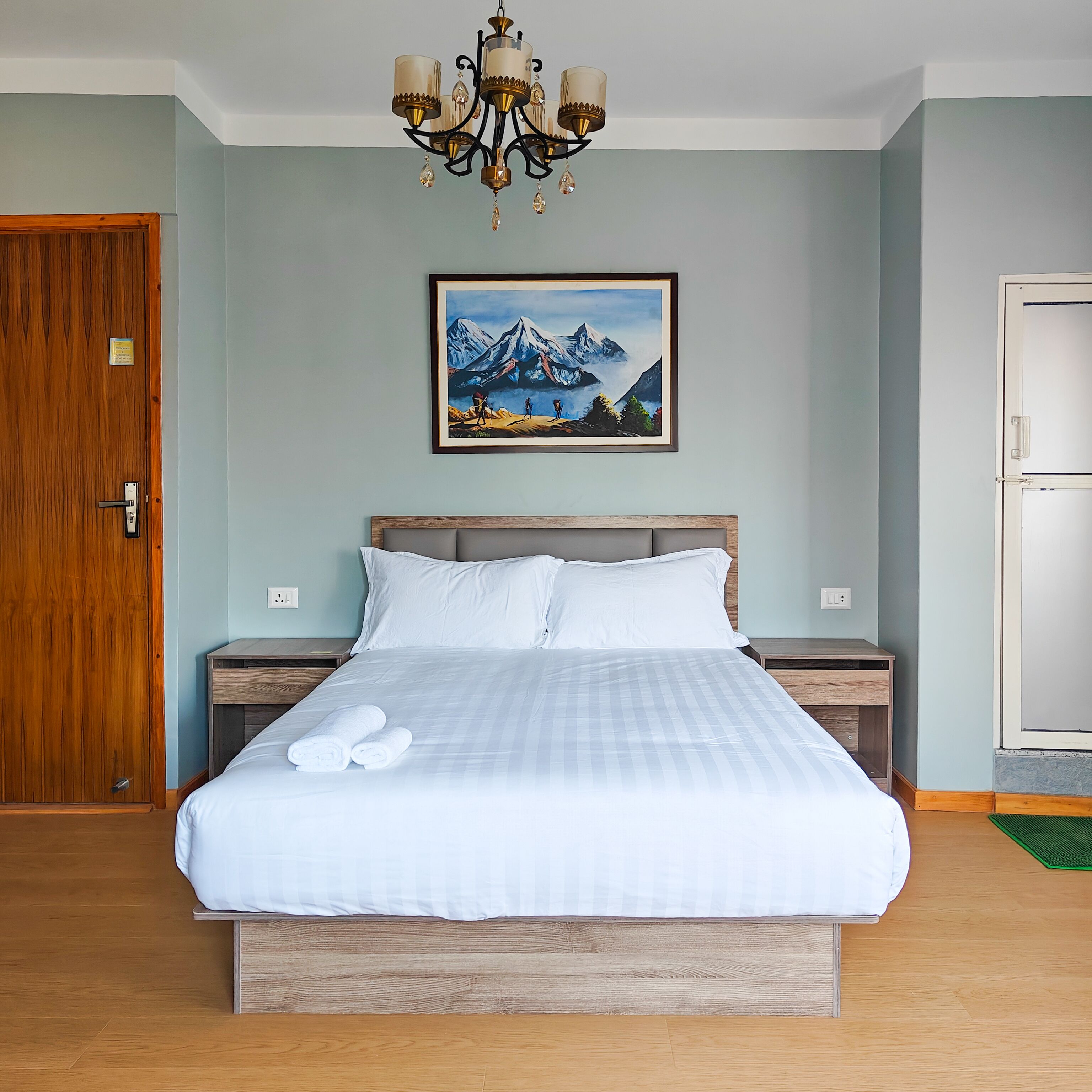 Ein Hotelzimmer mit einem großen Bett, einem hölzernen Kopfteil, einer Kristallleuchter, einem Gemälde von Bergen in einem Rahmen und einer Holztür.