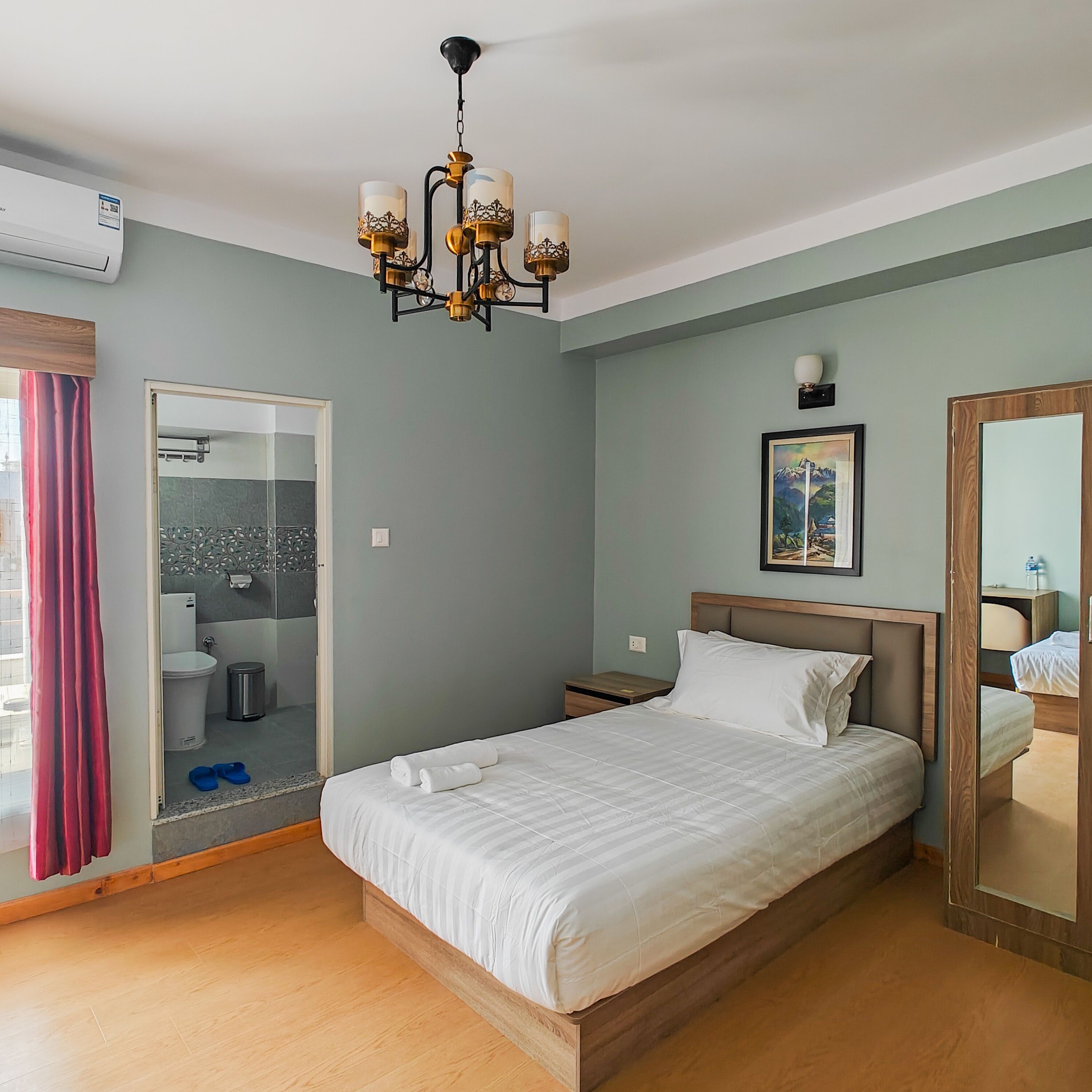A hotel room with a bed, a chandelier, a bathroom with a shower, and a painting on the wall.