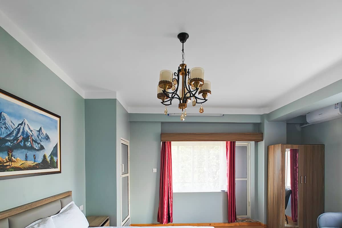 A hotel room with a large bed, a chandelier, a painting of mountains, a wardrobe, and a window with curtains.
