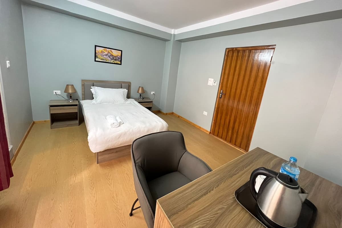 A hotel room with a bed, bedside tables, a desk with a kettle, a chair, and a wooden door.