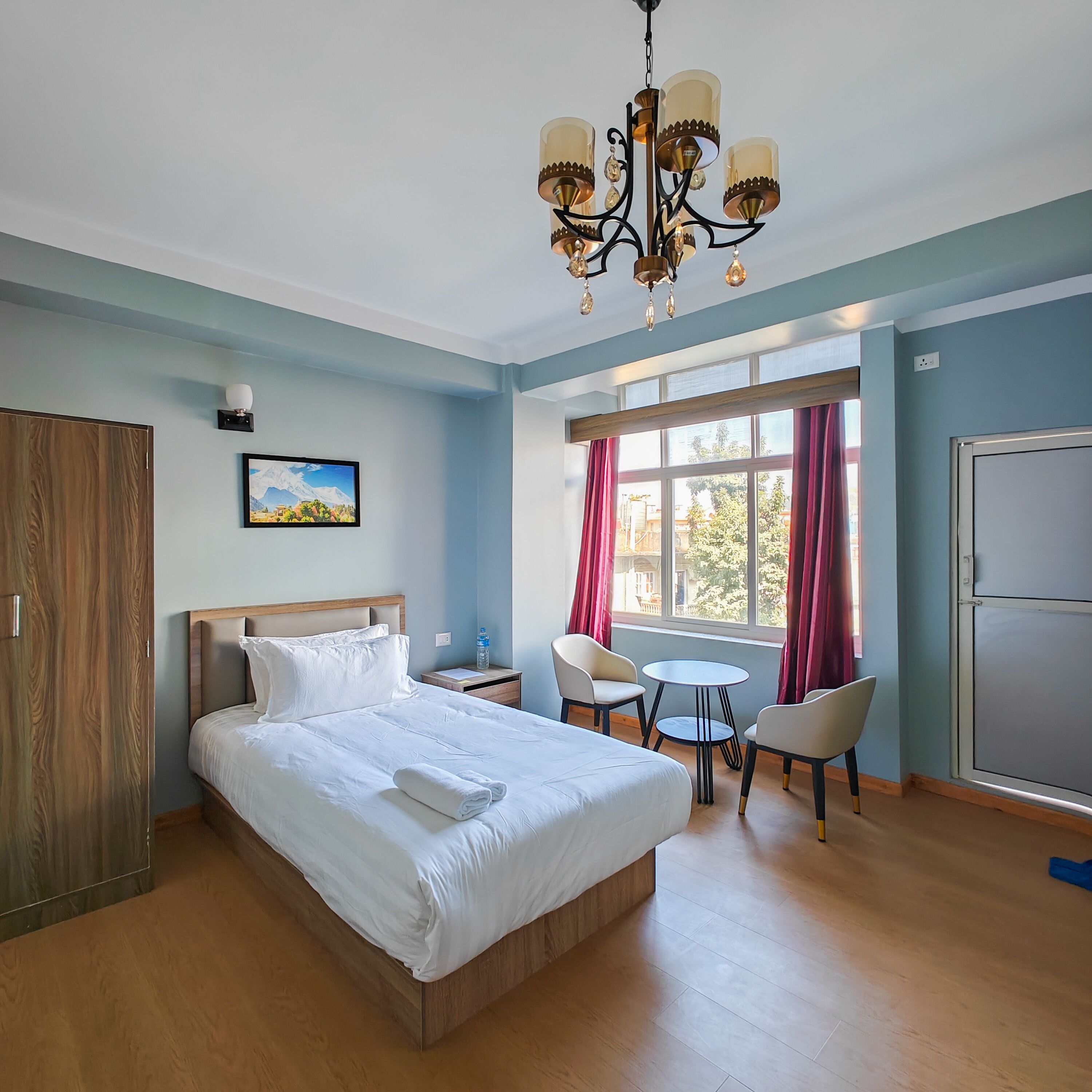 Hotellihuone, jossa on suuri sänky, työpöytä, kaksi tuolia, kattokruunu, ikkuna verhoilla ja vaatekaappi.