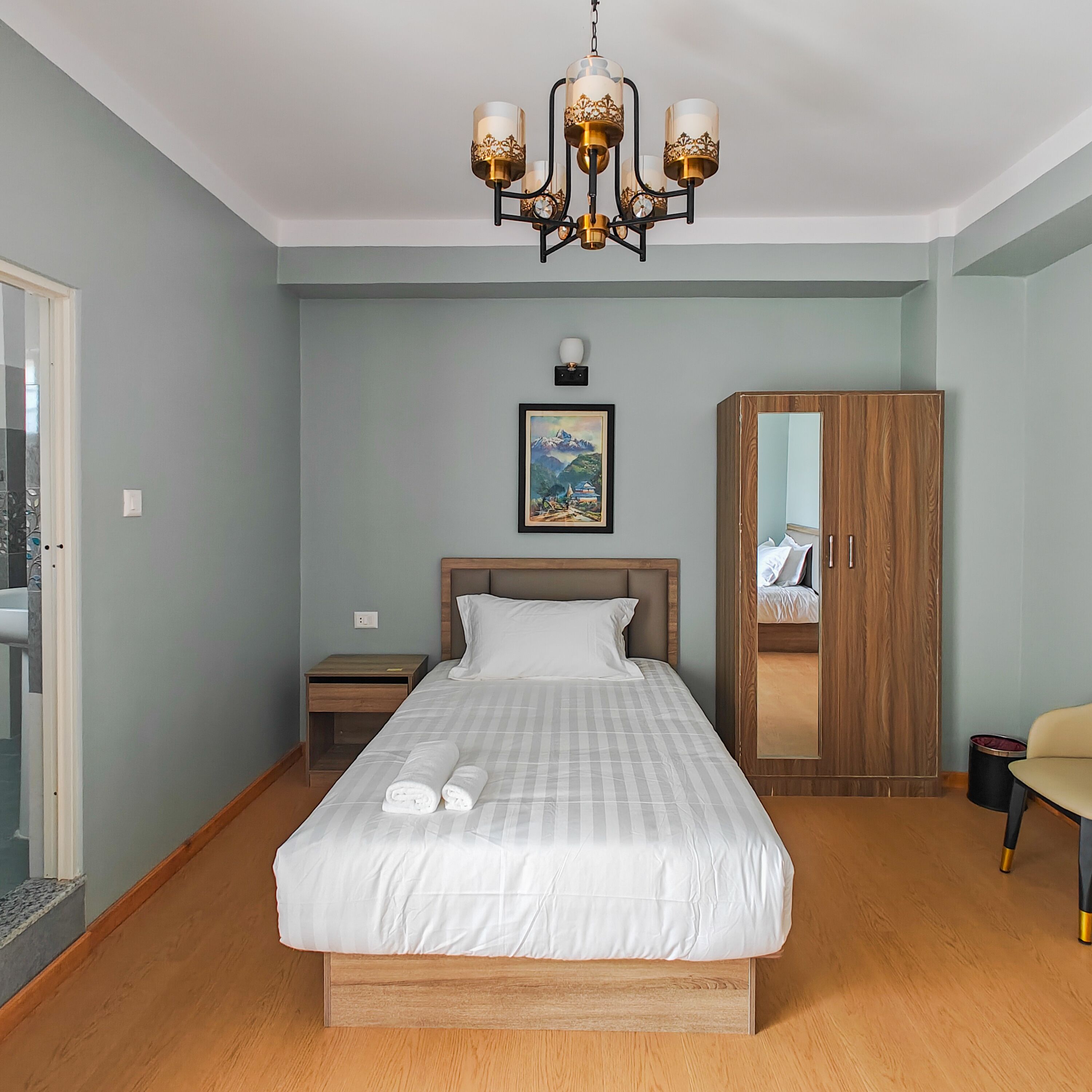 A bedroom with a bed, a wooden wardrobe, a nightstand, a chair, and a chandelier.