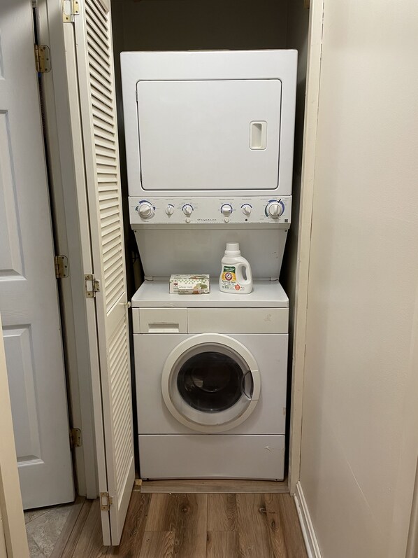 A compact laundry room with a front-loading washer and dryer, a small shelf with detergent bottles, and a door with a louvered window. - Cozy Long Pond Cottage: community access, private mooring & shared dock. (Rome)