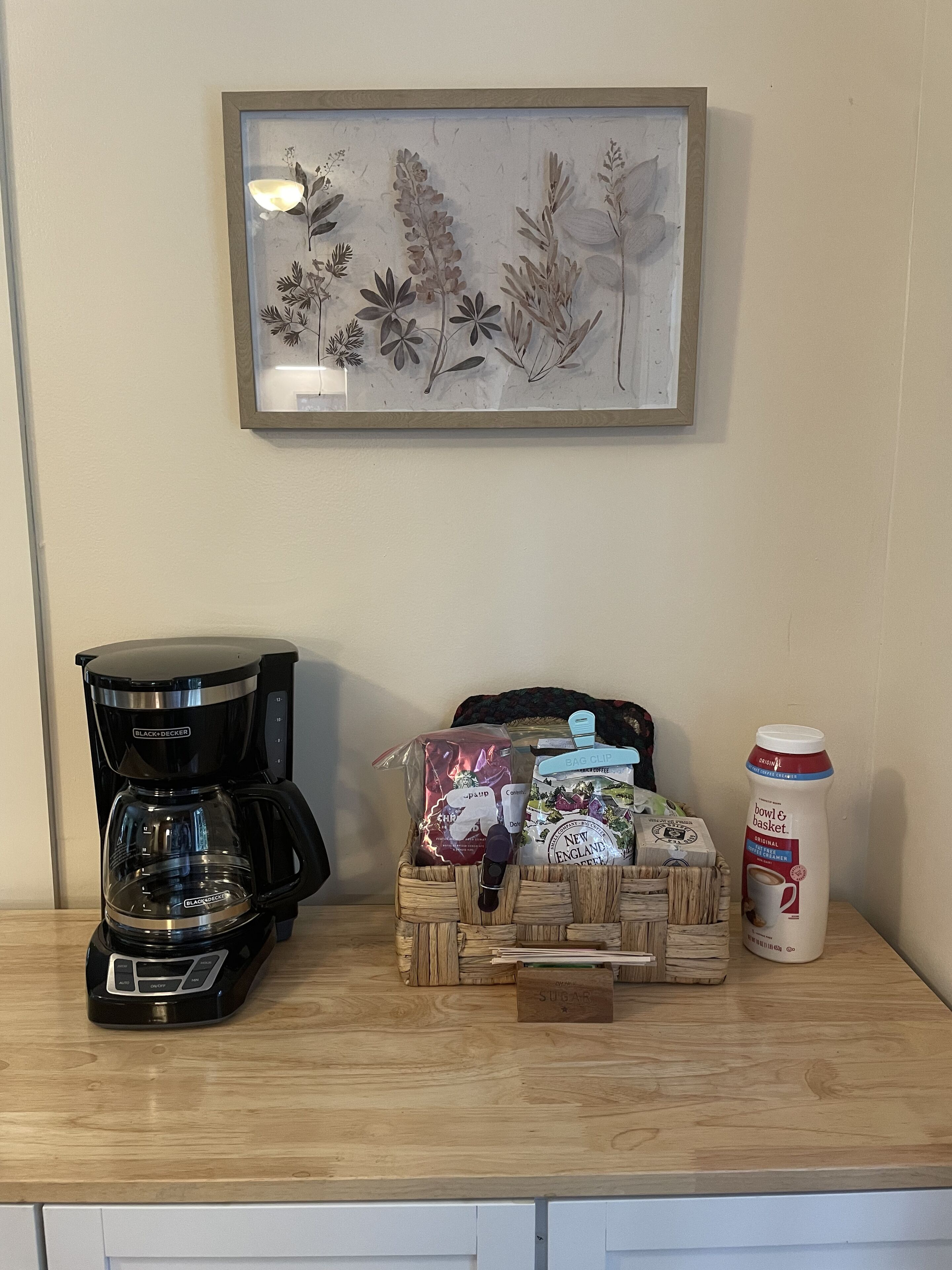 A coffee maker, a framed botanical print, a basket with coffee pods, a bag of sugar, and a milk carton on a wooden table.