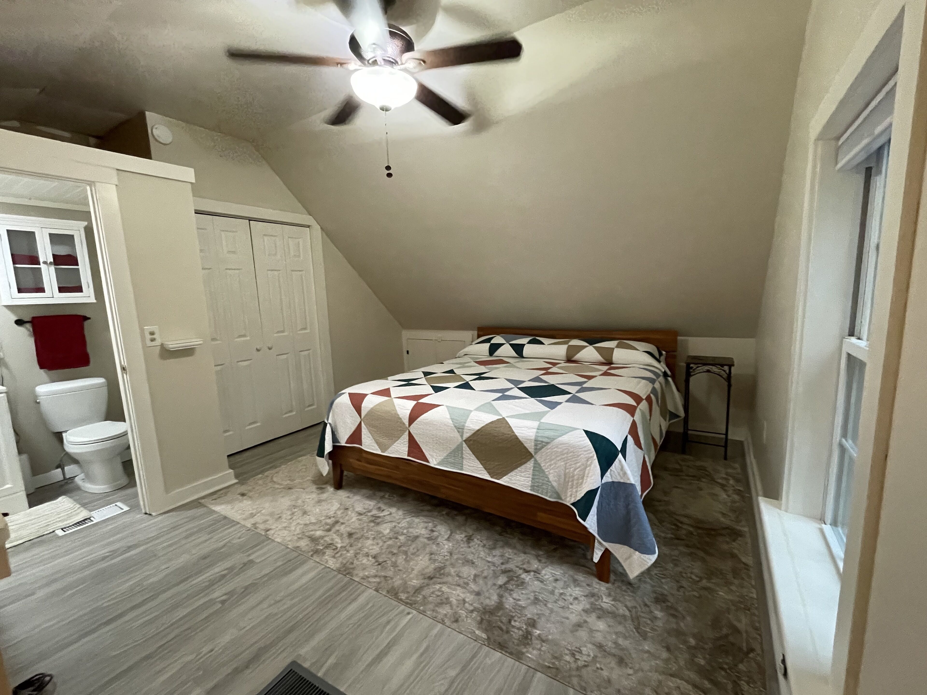 A bedroom with a sloped ceiling, a bed with a patterned quilt, a ceiling fan, and a window.