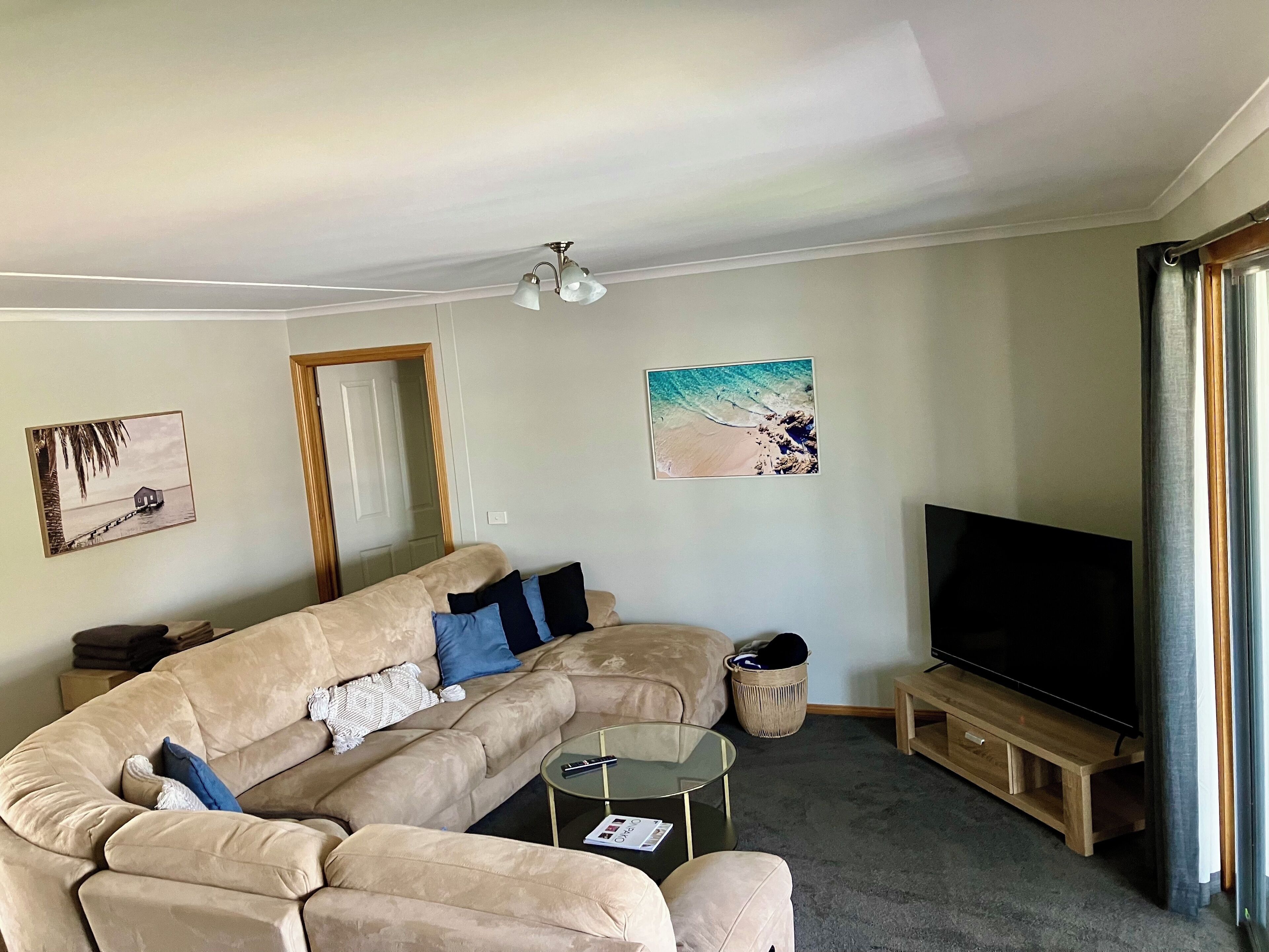 A living room with a beige sectional sofa, a glass coffee table, a TV on a wooden stand, and two wall-mounted pictures.