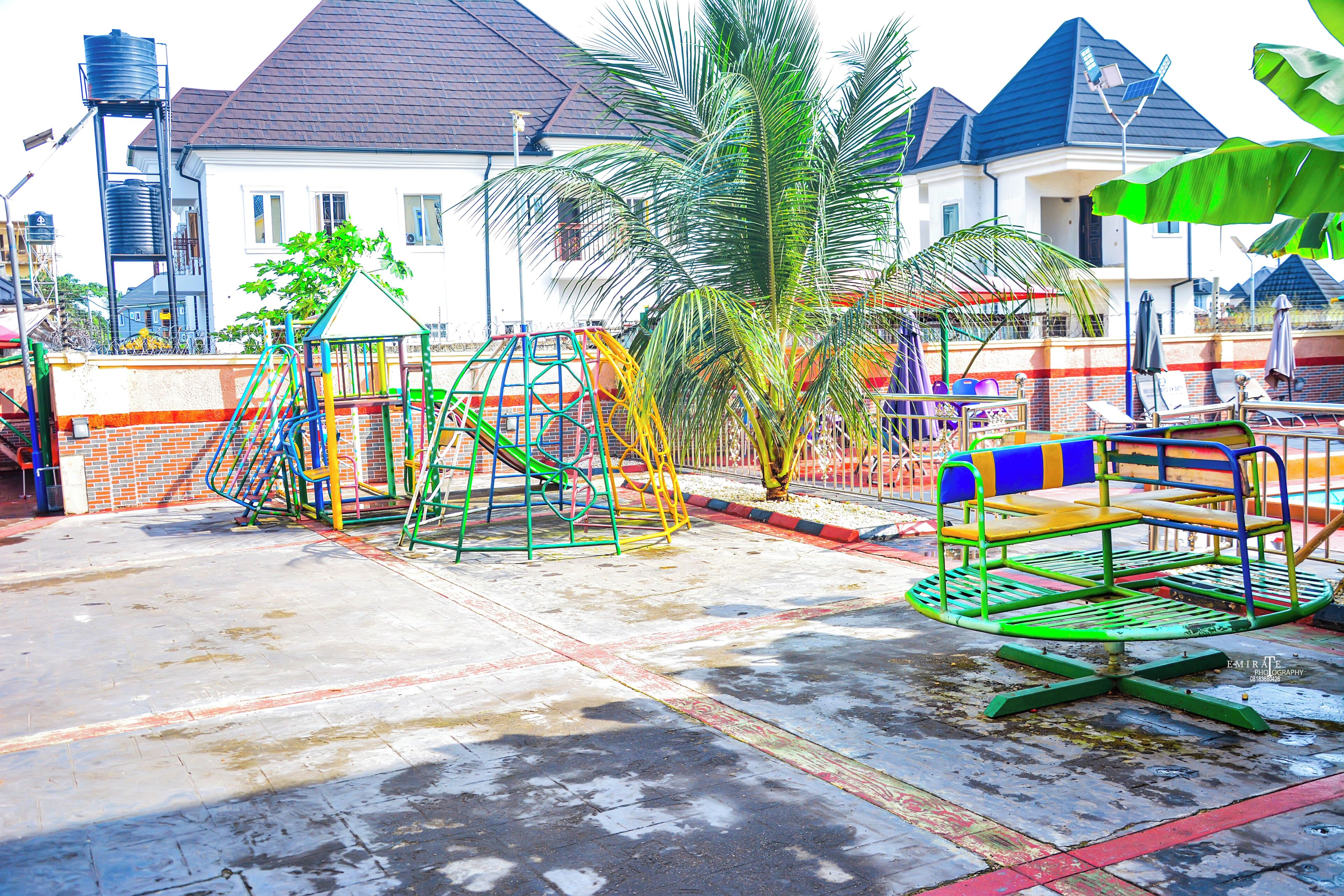 A playground with swings and climbing structures, a palm tree, and a house with a water tank in the background.