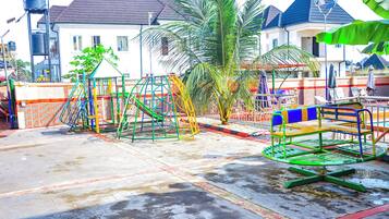 A playground with swings and climbing structures, a palm tree, and a house with a water tank in the background.